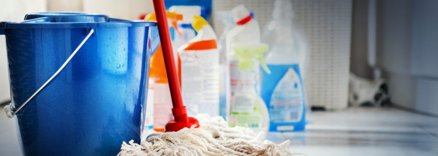 Closeup of unrdcognizable home cleaning products with blue bucket and a mop in front in sharp focus. All products placed on white and poorly lit bathroom floor.